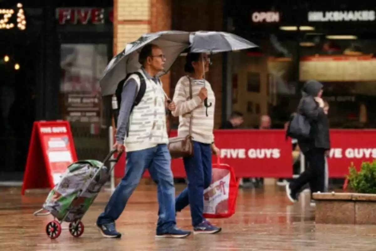 Dues persones caminen sota la pluja amb paraigües i porten bosses de compra davant d’un restaurant Five Guys.