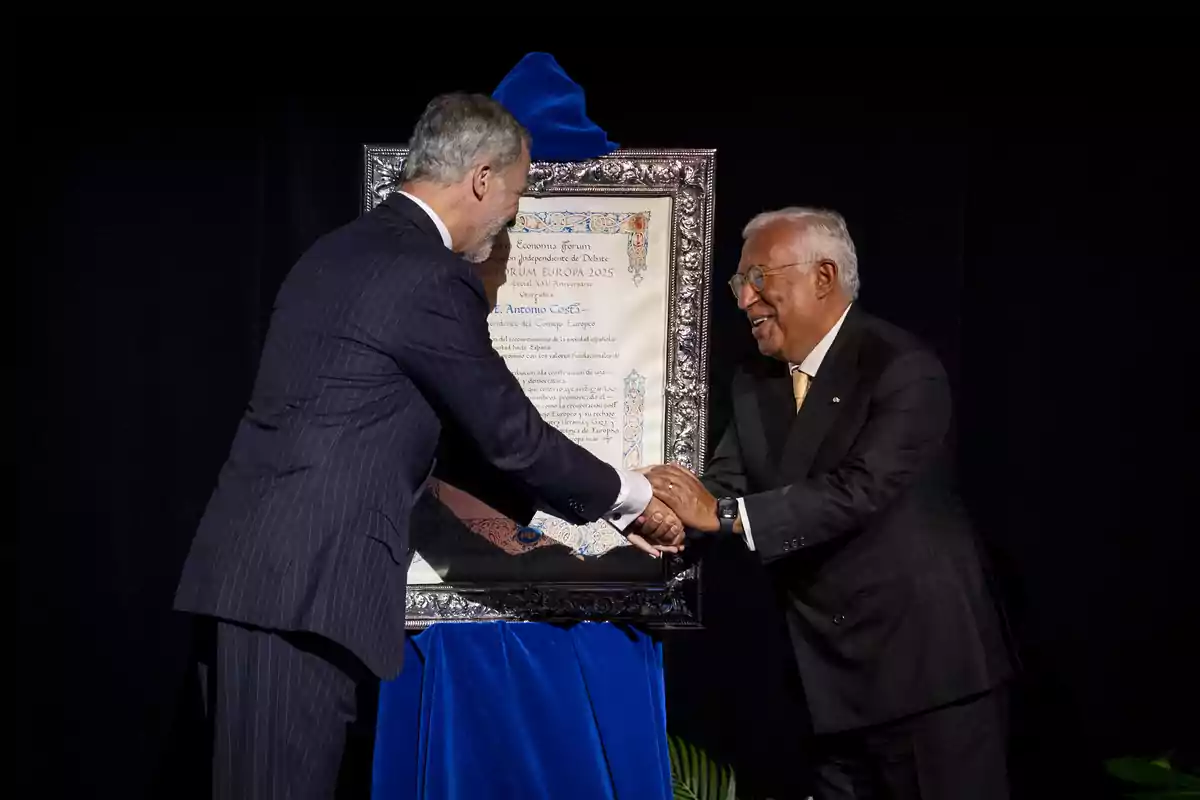 Felipe VI y Josep Borrell elegantemente vestidos se dan la mano sonrientes frente a un diploma enmarcado sobre un pedestal cubierto con tela azul Felipe VI y Josep Borrell elegantemente vestidos se dan la mano sonrientes frente a un diploma enmarcado sobre un pedestal cubierto con tela azul