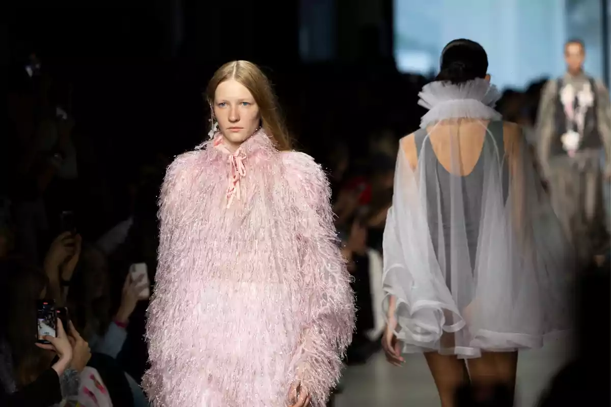 Desfile de moda con una modelo usando un vestido rosa de textura esponjosa y otra modelo de espaldas con un vestido gris y tul blanco Desfile de moda con una modelo usando un vestido rosa de textura esponjosa y otra modelo de espaldas con un vestido gris y tul blanco