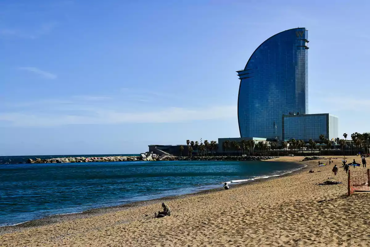 Playa de arena con personas disfrutando del sol y el mar junto a un edificio moderno de cristal en forma de vela en el fondo