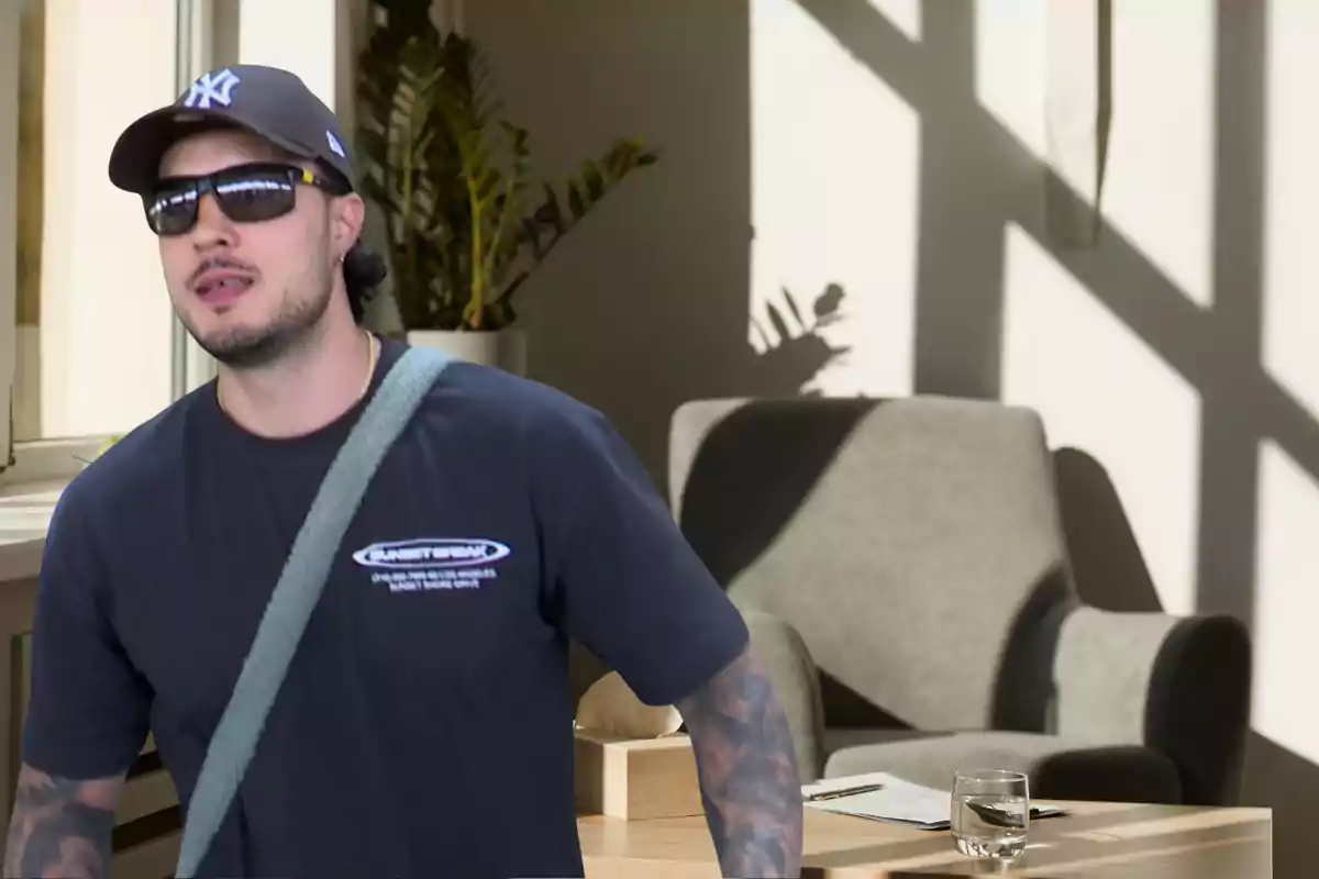 Carlo Costanzia con gorra y gafas de sol en una habitación iluminada con una silla gris y una mesa con un vaso de agua Carlo Costanzia con gorra y gafas de sol en una habitación iluminada con una silla gris y una mesa con un vaso de agua