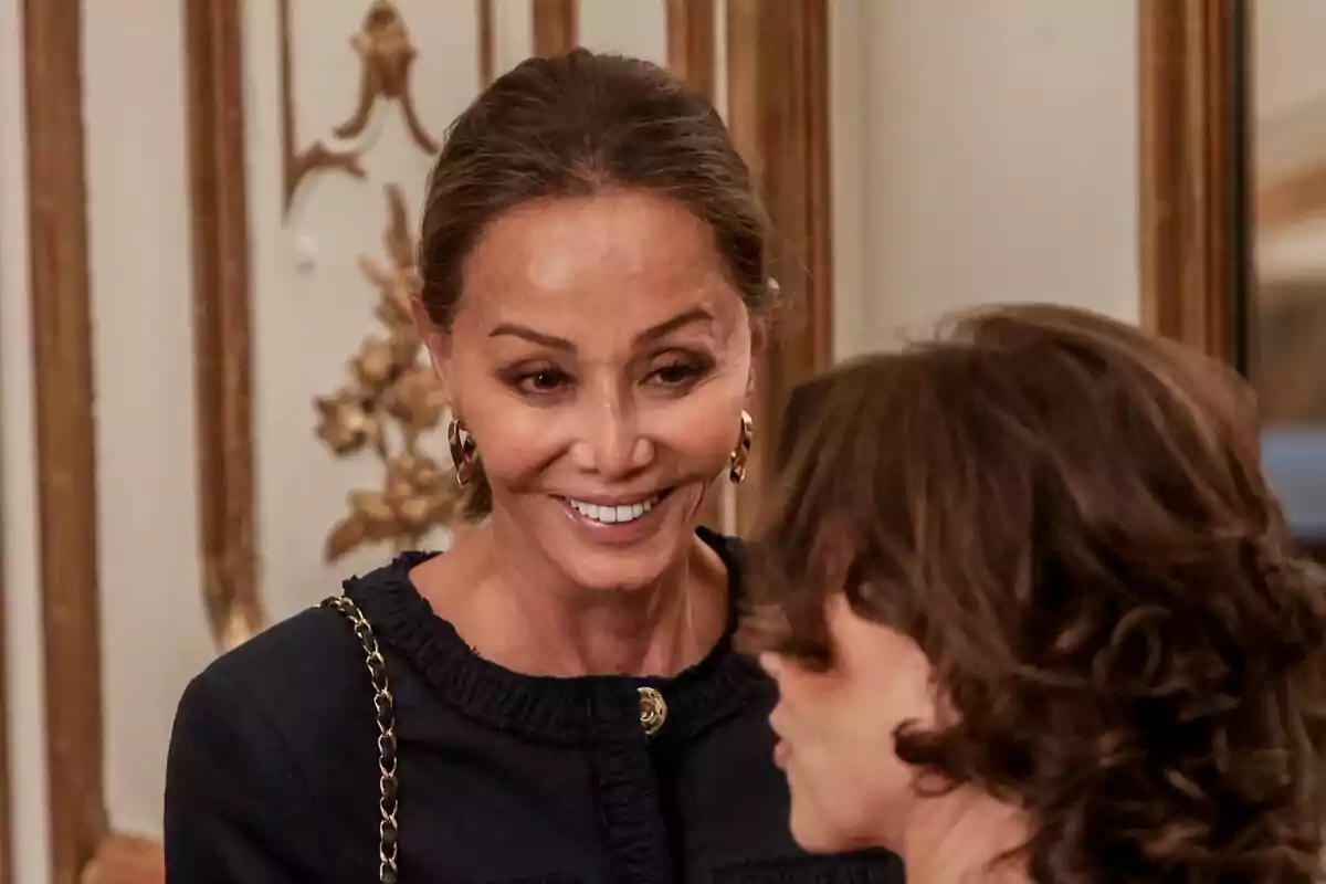 Dona somrient amb els cabells recollits i vestit fosc conversant amb una altra persona en un entorn elegant amb detalls daurats a la paret