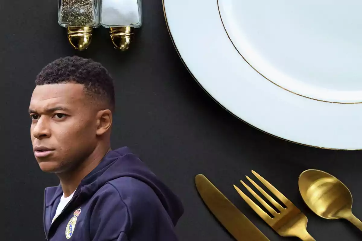 Un home amb jaqueta blau marí apareix davant d’un plat blanc, coberts daurats i saler i pebrer sobre una taula negra. Un home amb jaqueta blau marí apareix davant d’un plat blanc, coberts daurats i saler i pebrer sobre una taula negra.