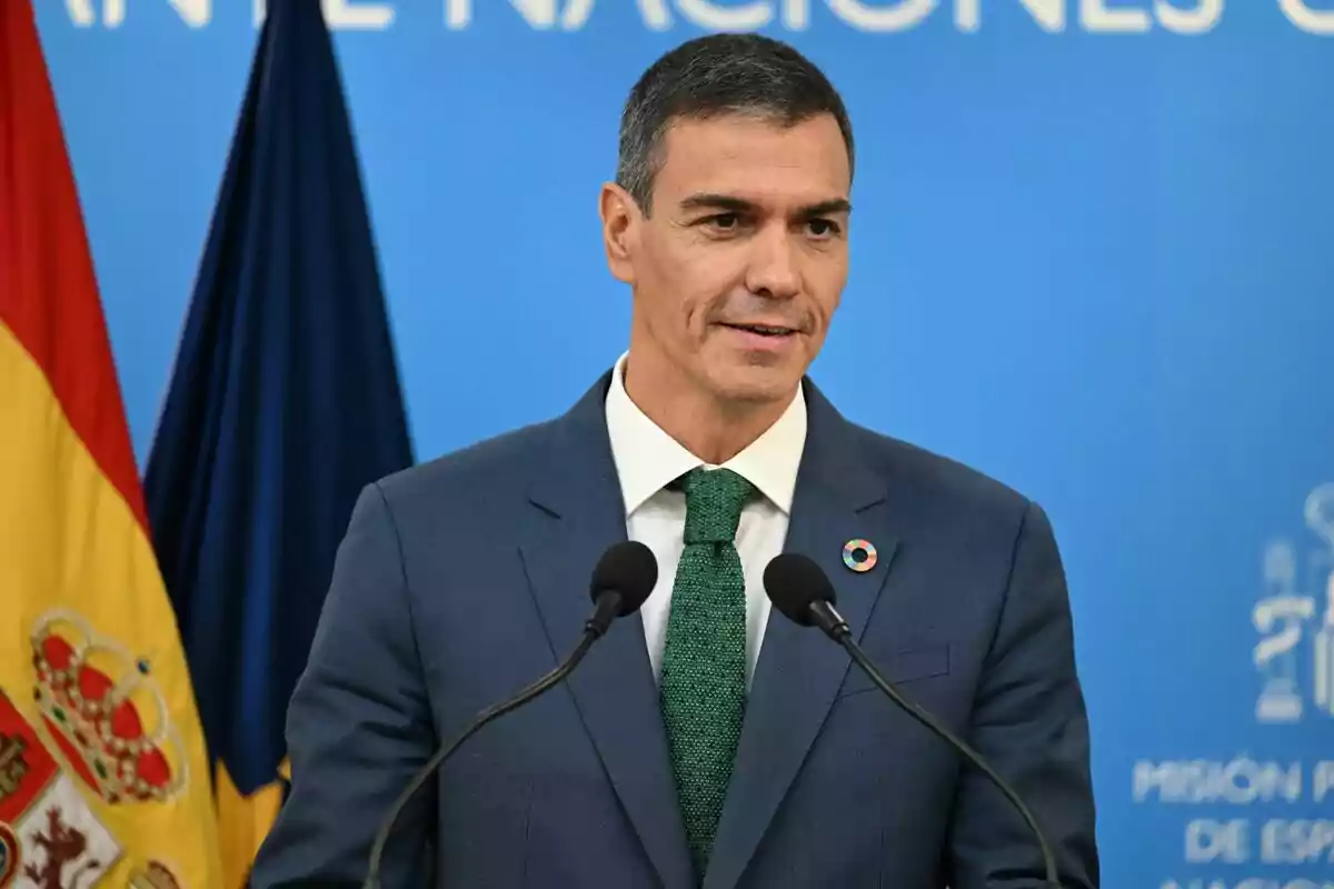 Home amb vestit blau i corbata verda parlant davant de micròfons amb una bandera d’Espanya al fons i un fons blau amb text blanc Home amb vestit blau i corbata verda parlant davant de micròfons amb una bandera d’Espanya al fons i un fons blau amb text blanc