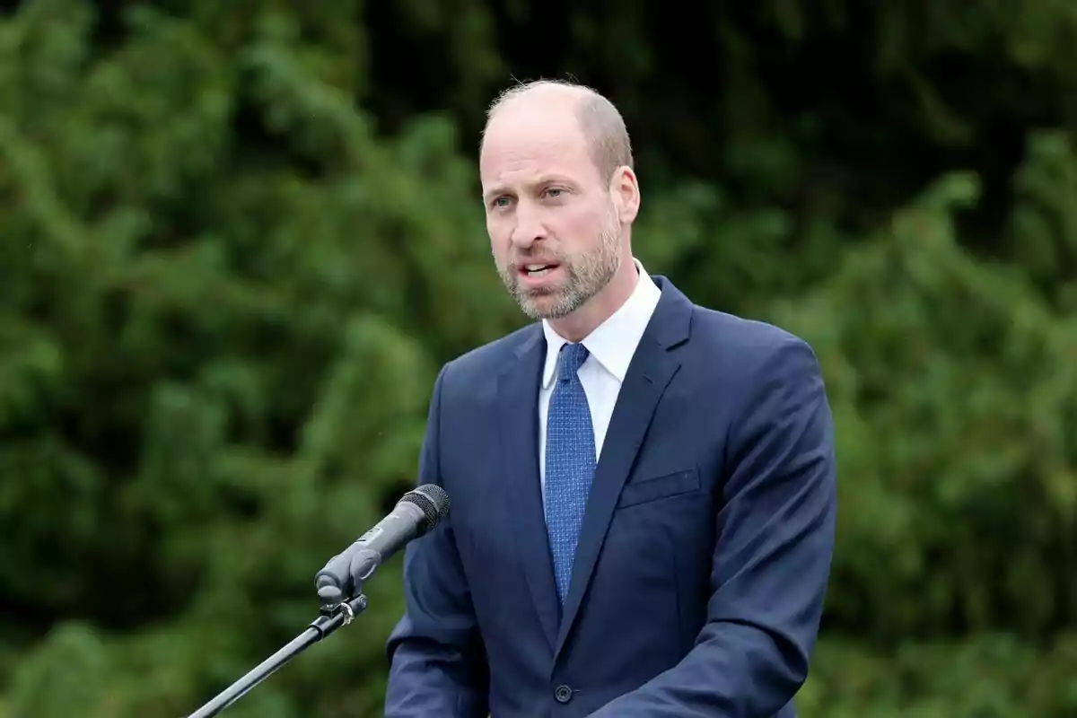Home amb barba i vestit blau parlant davant d’un micròfon a l’aire lliure amb un fons d’arbres verds Home amb barba i vestit blau parlant davant d’un micròfon a l’aire lliure amb un fons d’arbres verds