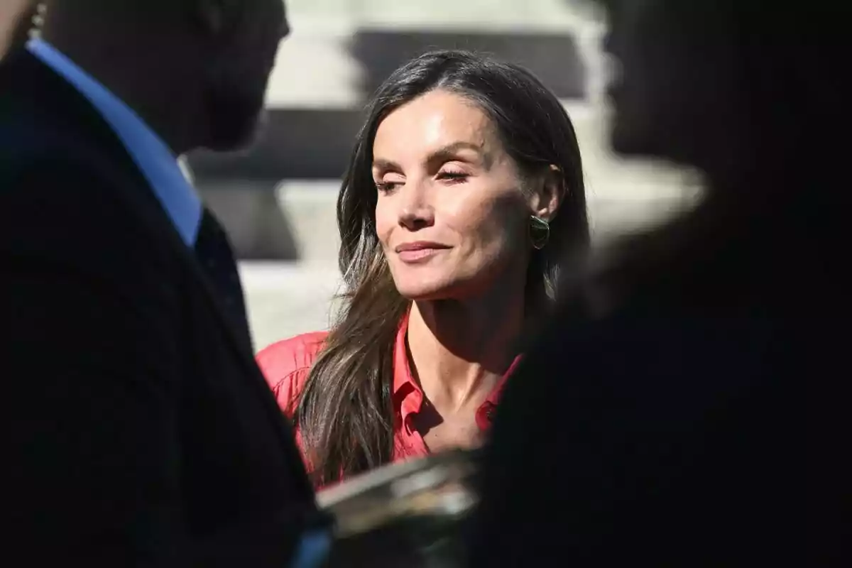 Mujer de cabello largo y oscuro con camisa roja y pendientes dorados, mirando hacia un lado mientras está parcialmente iluminada por la luz del sol, rodeada de otras personas desenfocadas. Mujer de cabello largo y oscuro con camisa roja y pendientes dorados, mirando hacia un lado mientras está parcialmente iluminada por la luz del sol, rodeada de otras personas desenfocadas.