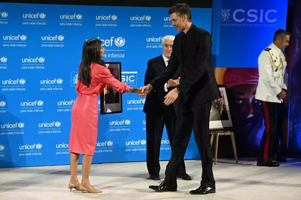 Una mujer con vestido rosa entrega un reconocimiento a un hombre alto vestido de negro en un evento de UNICEF mientras otras dos personas observan en el escenario Una mujer con vestido rosa entrega un reconocimiento a un hombre alto vestido de negro en un evento de UNICEF mientras otras dos personas observan en el escenario