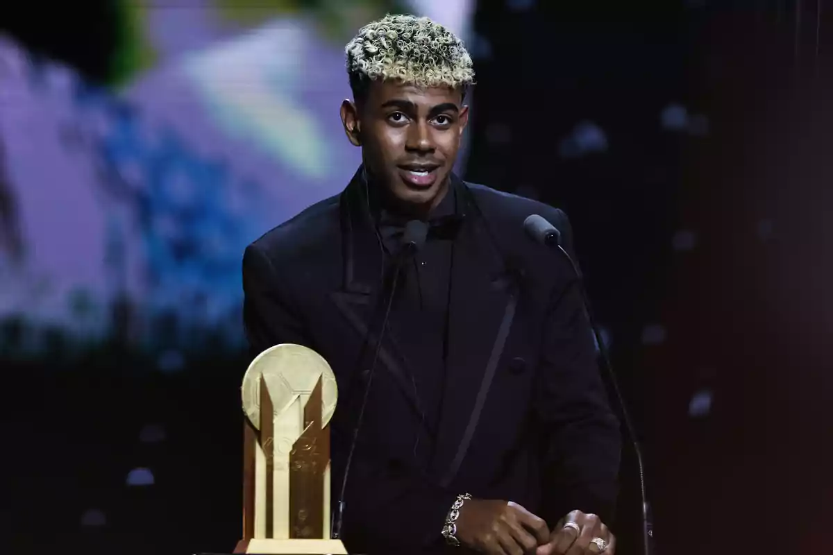 Lamine Yamal amb un vestit negre, dret davant d’un micròfon i un trofeu daurat sobre una taula, en un escenari il·luminat Lamine Yamal amb un vestit negre, dret davant d’un micròfon i un trofeu daurat sobre una taula, en un escenari il·luminat