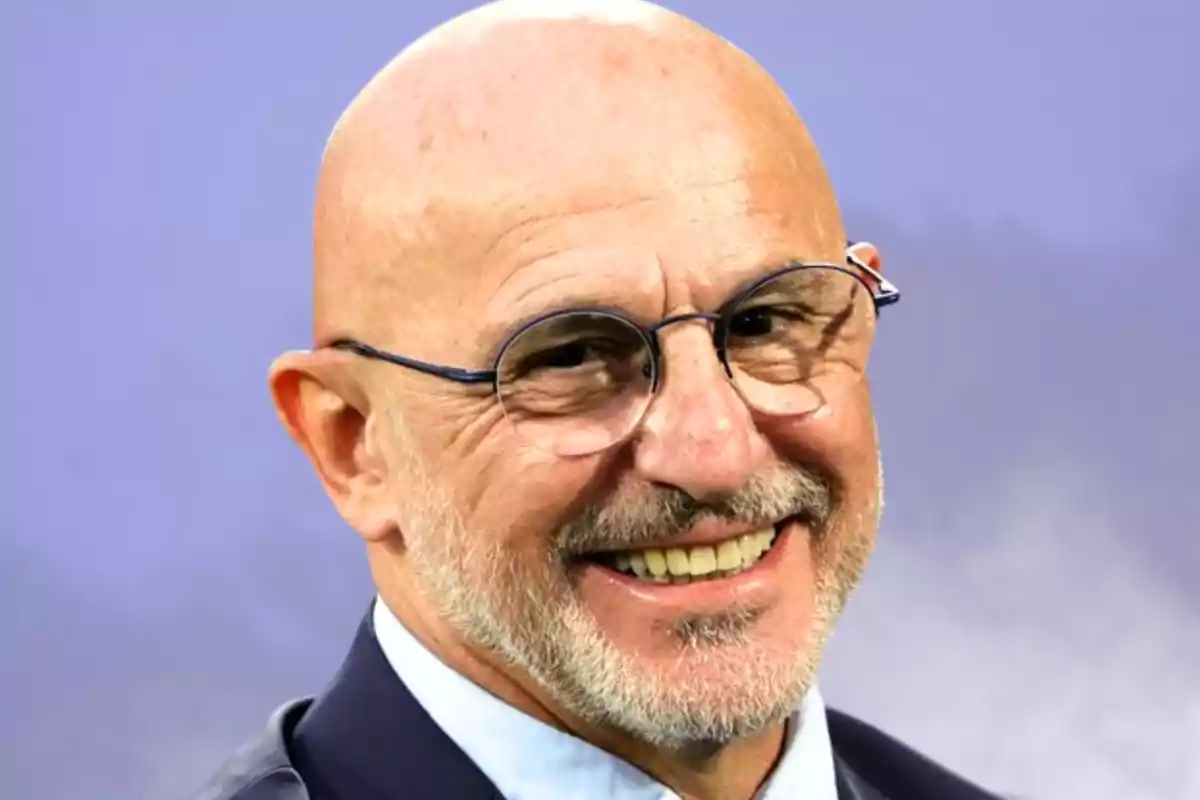 Hombre calvo con gafas y barba sonriente usando saco y camisa clara frente a un fondo lila difuminado