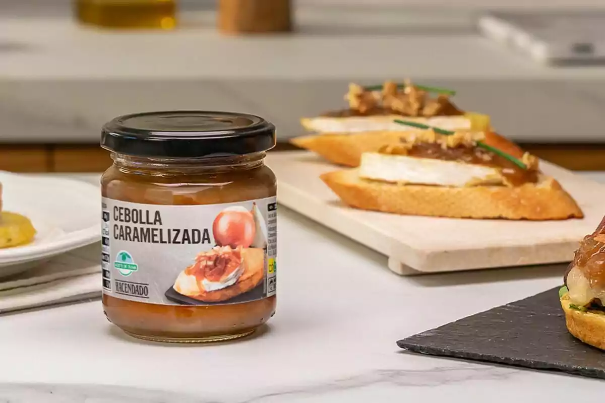 Un frasco de cebolla caramelizada sobre una mesa junto a unas tostadas con queso y cebolla caramelizada como aperitivo Un frasco de cebolla caramelizada sobre una mesa junto a unas tostadas con queso y cebolla caramelizada como aperitivo