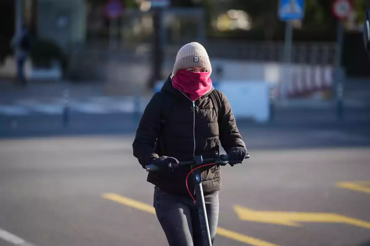 Persona abrigada con gorro y bufanda rosa conduciendo un patinete eléctrico por la ciudad en un día frío.