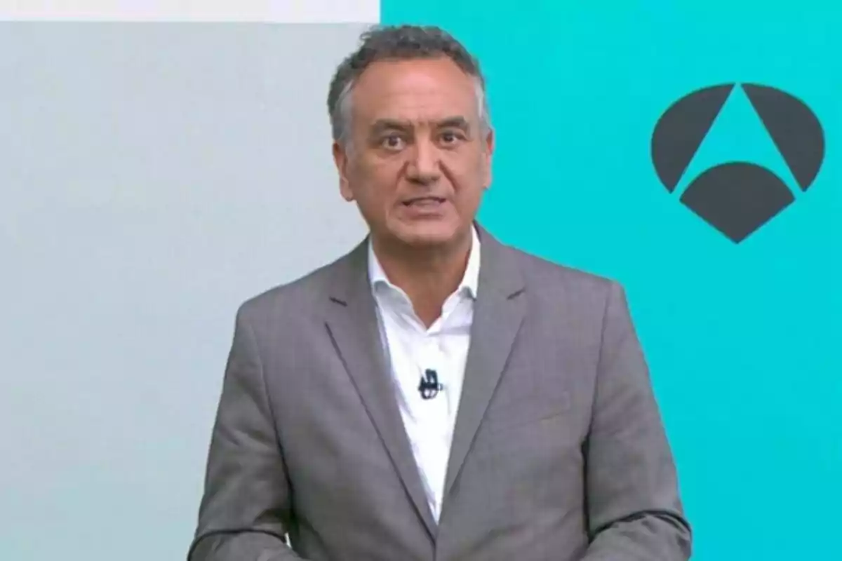 Roberto Brasero vestit amb vestit gris i camisa blanca dret davant d’un fons blau amb un logotip d’Antena 3.