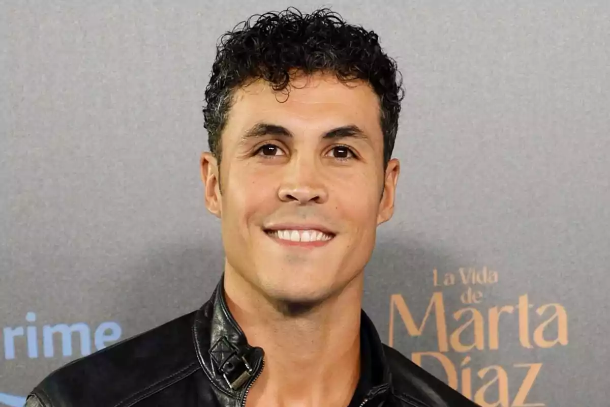 Kiko Jiménez de cabello rizado y corto sonriendo, vestido con chaqueta de cuero negra, posando frente a un fondo gris con letras desenfocadas