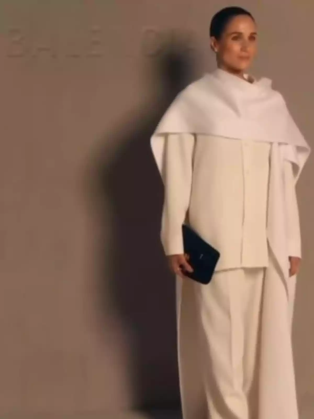 Meghan Markle de pie vestida con un conjunto elegante de color blanco y sosteniendo un bolso negro Meghan Markle de pie vestida con un conjunto elegante de color blanco y sosteniendo un bolso negro