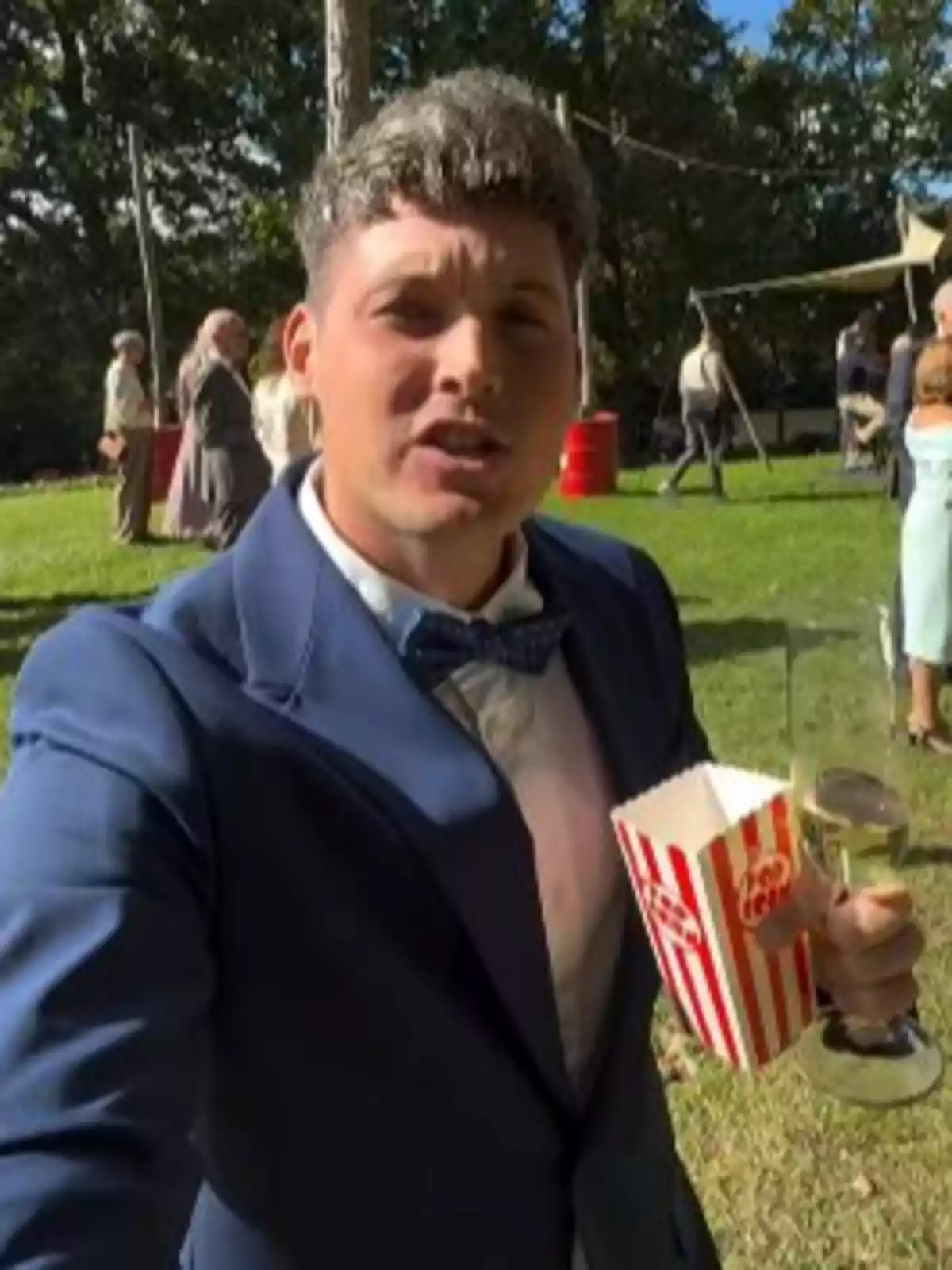 Home jove amb vestit blau sostenint una copa de vi i una capsa de crispetes en un esdeveniment a l’aire lliure amb diverses persones al fons Home jove amb vestit blau sostenint una copa de vi i una capsa de crispetes en un esdeveniment a l’aire lliure amb diverses persones al fons