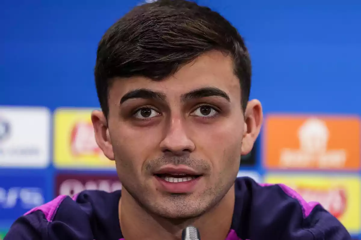 Pedri hablando frente a un micrófono con fondo azul y logotipos desenfocados Pedri hablando frente a un micrófono con fondo azul y logotipos desenfocados