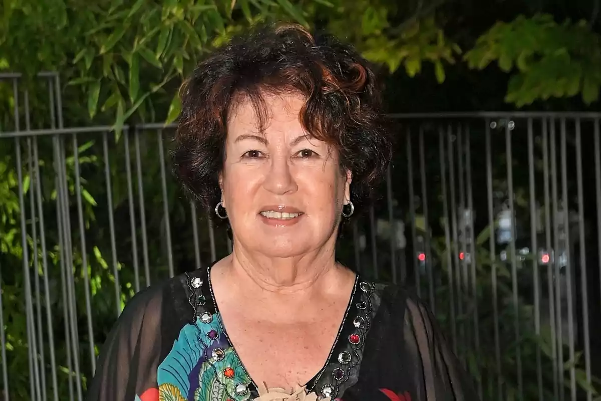 Rosa Villascastín, con cabello corto y rizado y blusa negra decorada, posando al aire libre frente a una reja y vegetación.