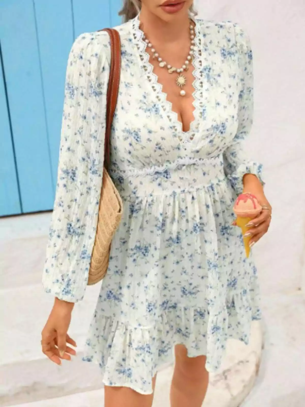 Dona amb vestit blanc de flors blaves, bossa de ràfia, collaret de perles i gelat a la mà davant d’una porta blava