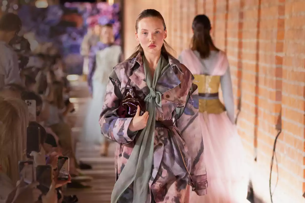 Modelo desfilando en una pasarela con un abrigo de estampado artístico y bufanda verde mientras el público toma fotos con sus teléfonos Modelo desfilando en una pasarela con un abrigo de estampado artístico y bufanda verde mientras el público toma fotos con sus teléfonos