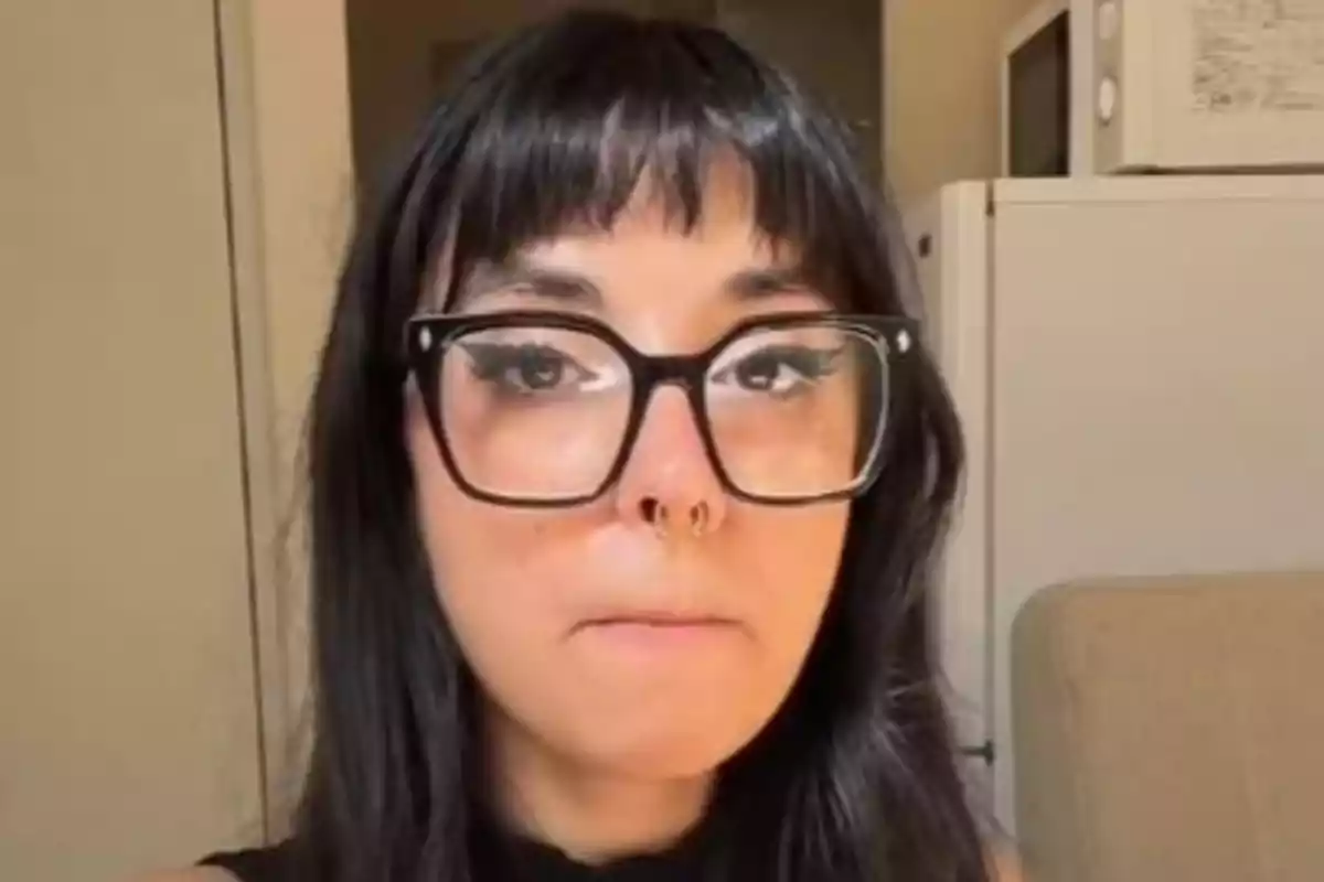 Dona de cabells foscos i ulleres grosses amb expressió seriosa en una habitació amb electrodomèstics al fons Dona de cabells foscos i ulleres grosses amb expressió seriosa en una habitació amb electrodomèstics al fons