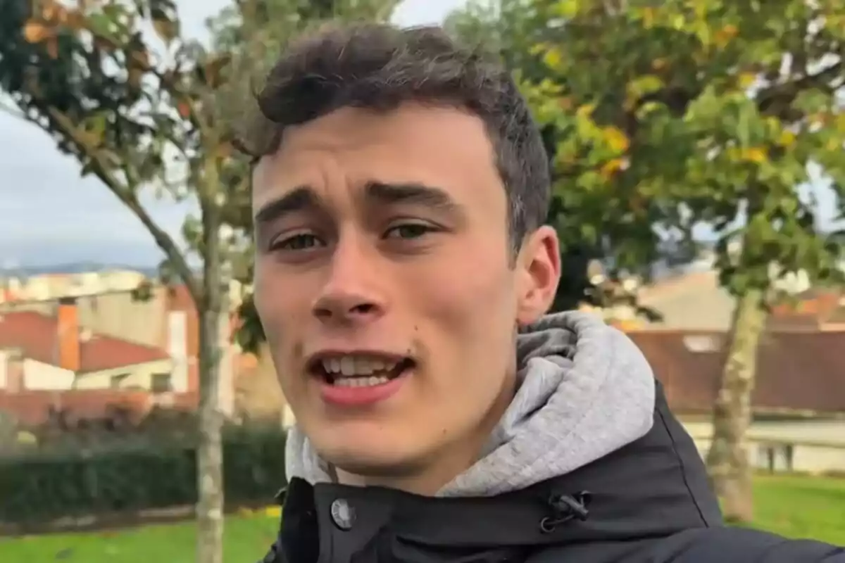 Jorge Rey vestido con chaqueta negra y sudadera gris posando al aire libre en un parque con árboles y casas al fondo. Jorge Rey vestido con chaqueta negra y sudadera gris posando al aire libre en un parque con árboles y casas al fondo.