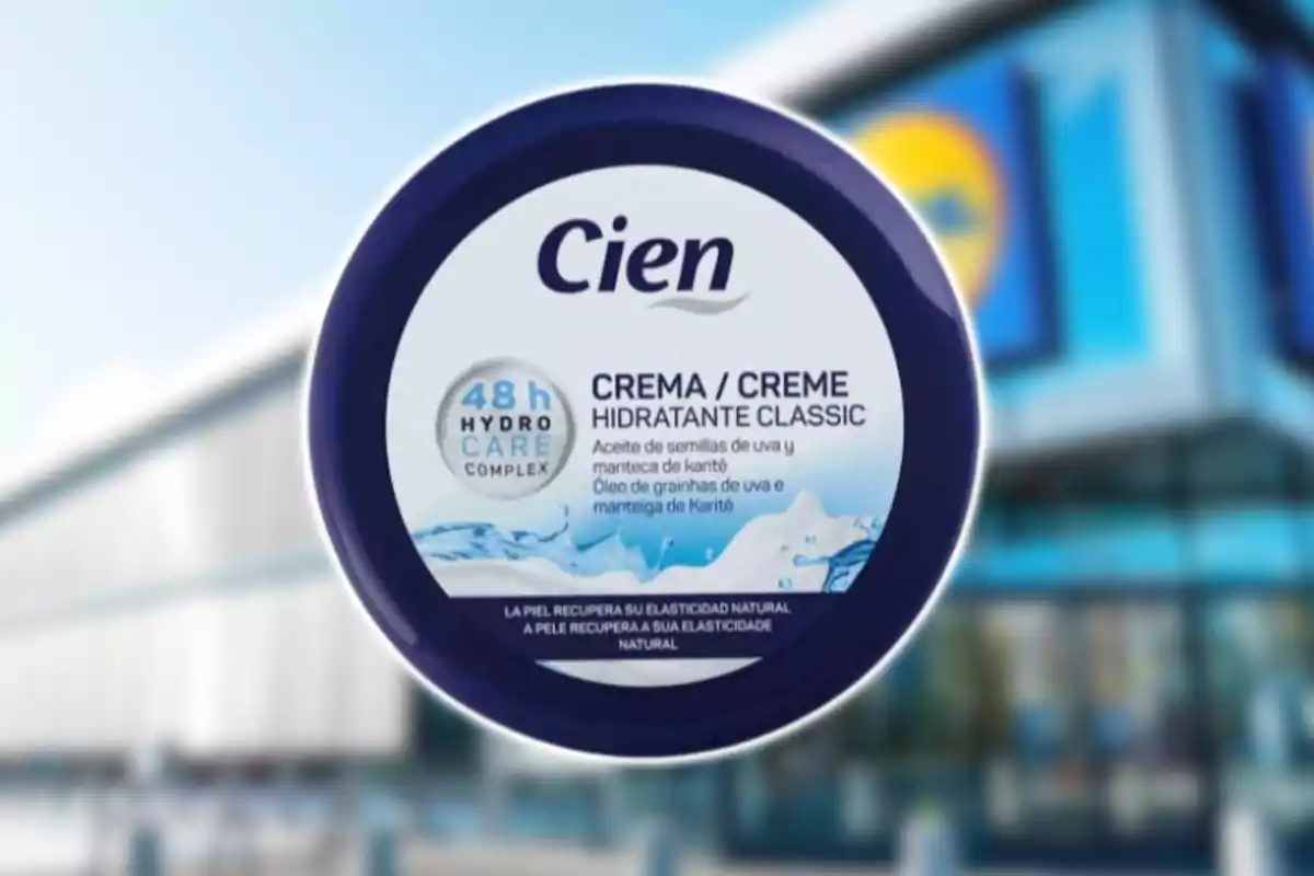 Envàs rodó de crema hidratant Cien amb tapa blava i etiqueta blanca, al fons una botiga amb façana blava desenfocada.