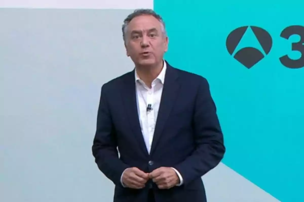 Roberto Brasero vestit amb vestit fosc i camisa blanca dret davant d’un fons blau clar amb un logotip d’Antena 3.