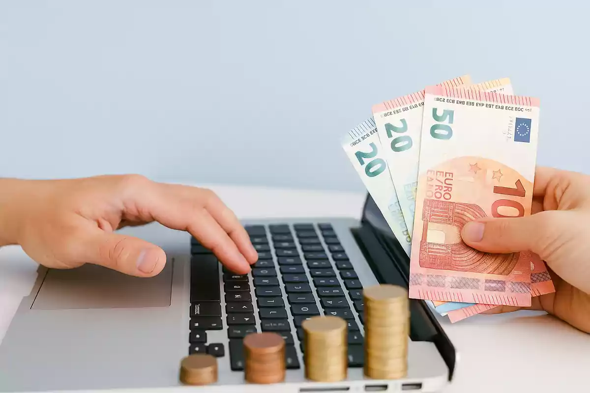 Persona usando una computadora portátil con una mano mientras sostiene billetes de euro y hay varias pilas de monedas sobre el teclado