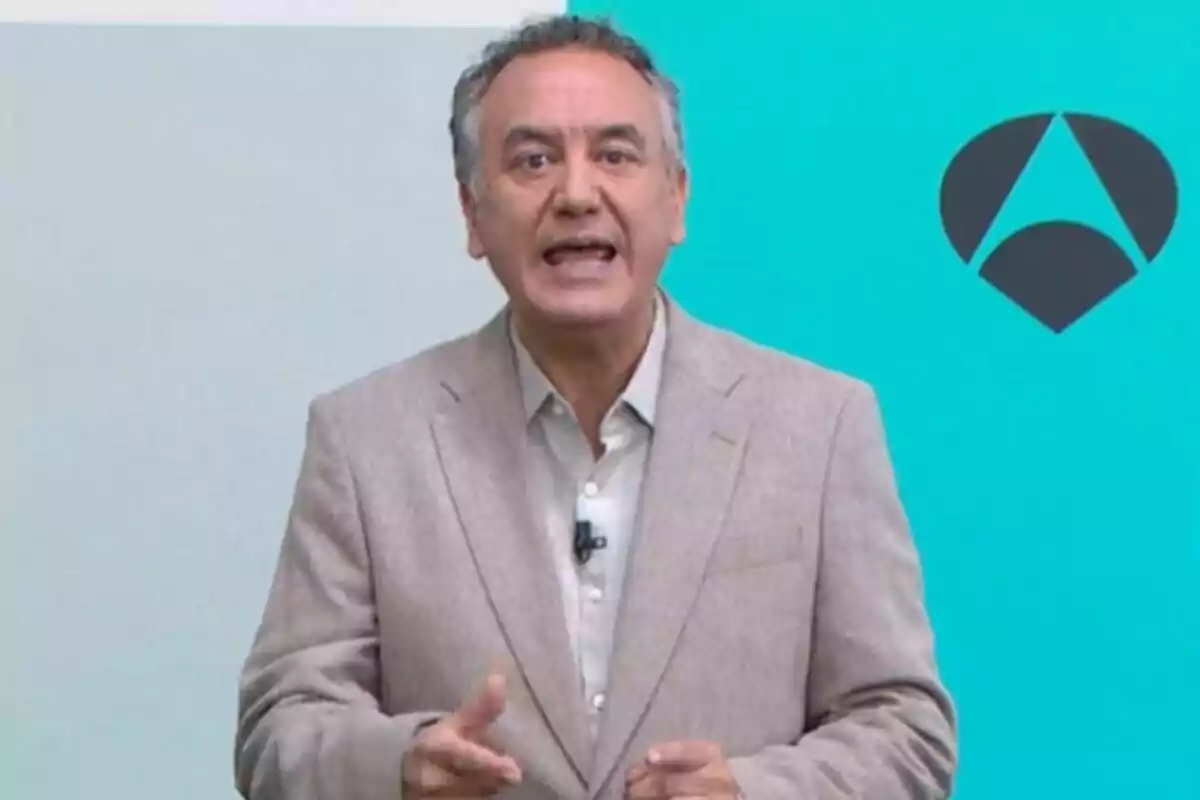 Roberto Brasero vestit amb un vestit beix parlant davant d’un fons blau amb un logotip geomètric negre, que és d’Antena 3.