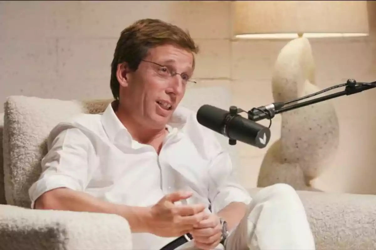 José Luis Martínez-Almeida con camisa blanca hablando en un micrófono mientras está sentado en un sillón con una lámpara de fondo. José Luis Martínez-Almeida con camisa blanca hablando en un micrófono mientras está sentado en un sillón con una lámpara de fondo.