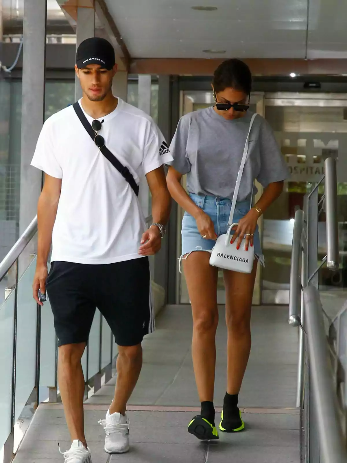 Achraf Hakimi y Hiba Abouk caminando por un pasillo techado, él lleva una camiseta blanca y pantalones cortos negros, mientras que ella viste una camiseta gris y shorts de mezclilla. Achraf Hakimi y Hiba Abouk caminando por un pasillo techado, él lleva una camiseta blanca y pantalones cortos negros, mientras que ella viste una camiseta gris y shorts de mezclilla.
