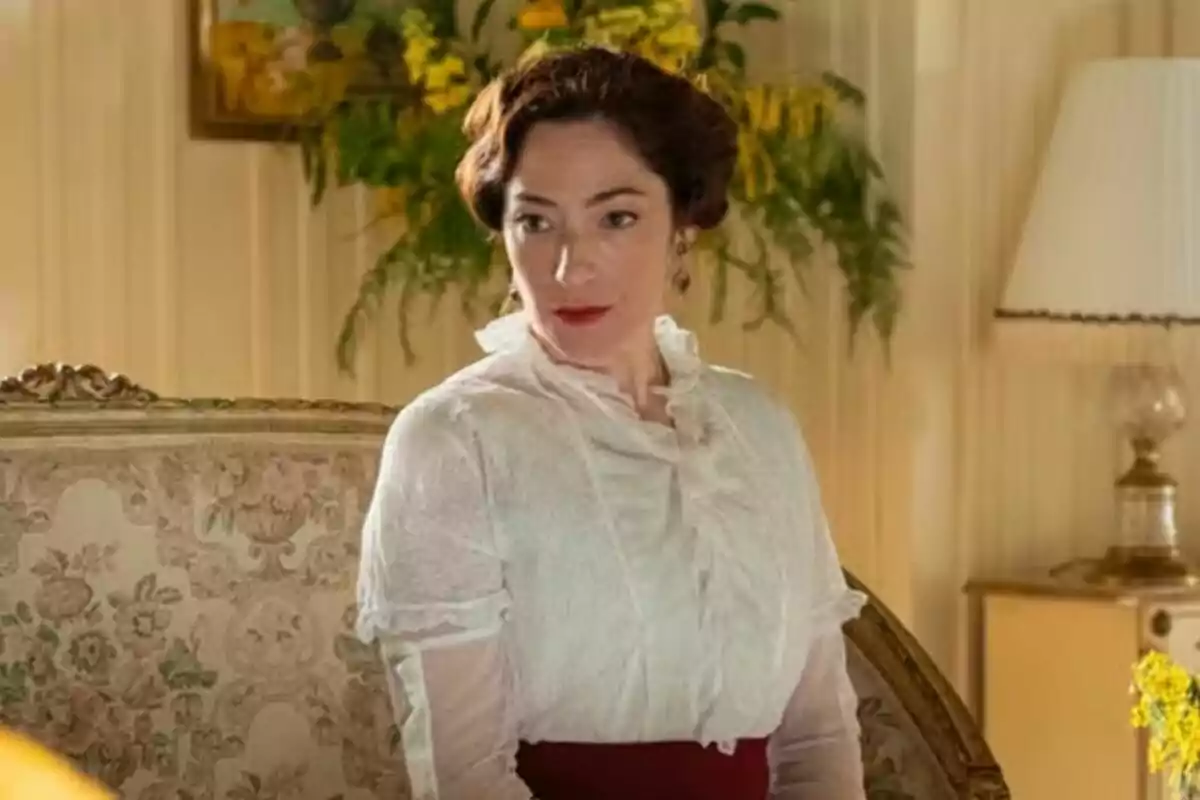 L'actriu Alicia Moruno vestida amb brusa blanca i faldilla vermella està asseguda en un sofà decorat amb flors en una habitació elegant amb una làmpada i plantes al fons. L'actriu Alicia Moruno vestida amb brusa blanca i faldilla vermella està asseguda en un sofà decorat amb flors en una habitació elegant amb una làmpada i plantes al fons.