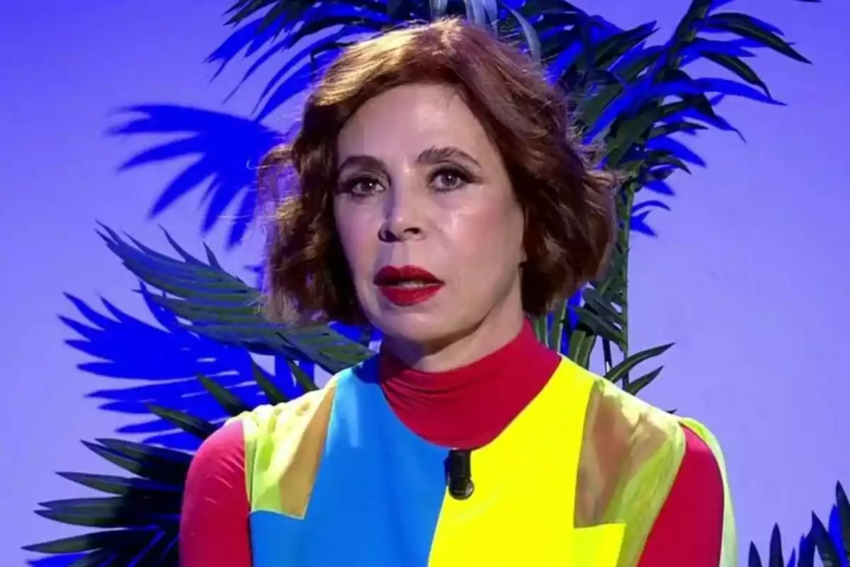 Ágatha Ruiz de la Prada amb cabell curt i castany, vestida amb un vestit acolorit, està davant d'un fons blau amb fulles de plantes a De Viernes. Ágatha Ruiz de la Prada amb cabell curt i castany, vestida amb un vestit acolorit, està davant d'un fons blau amb fulles de plantes a De Viernes.