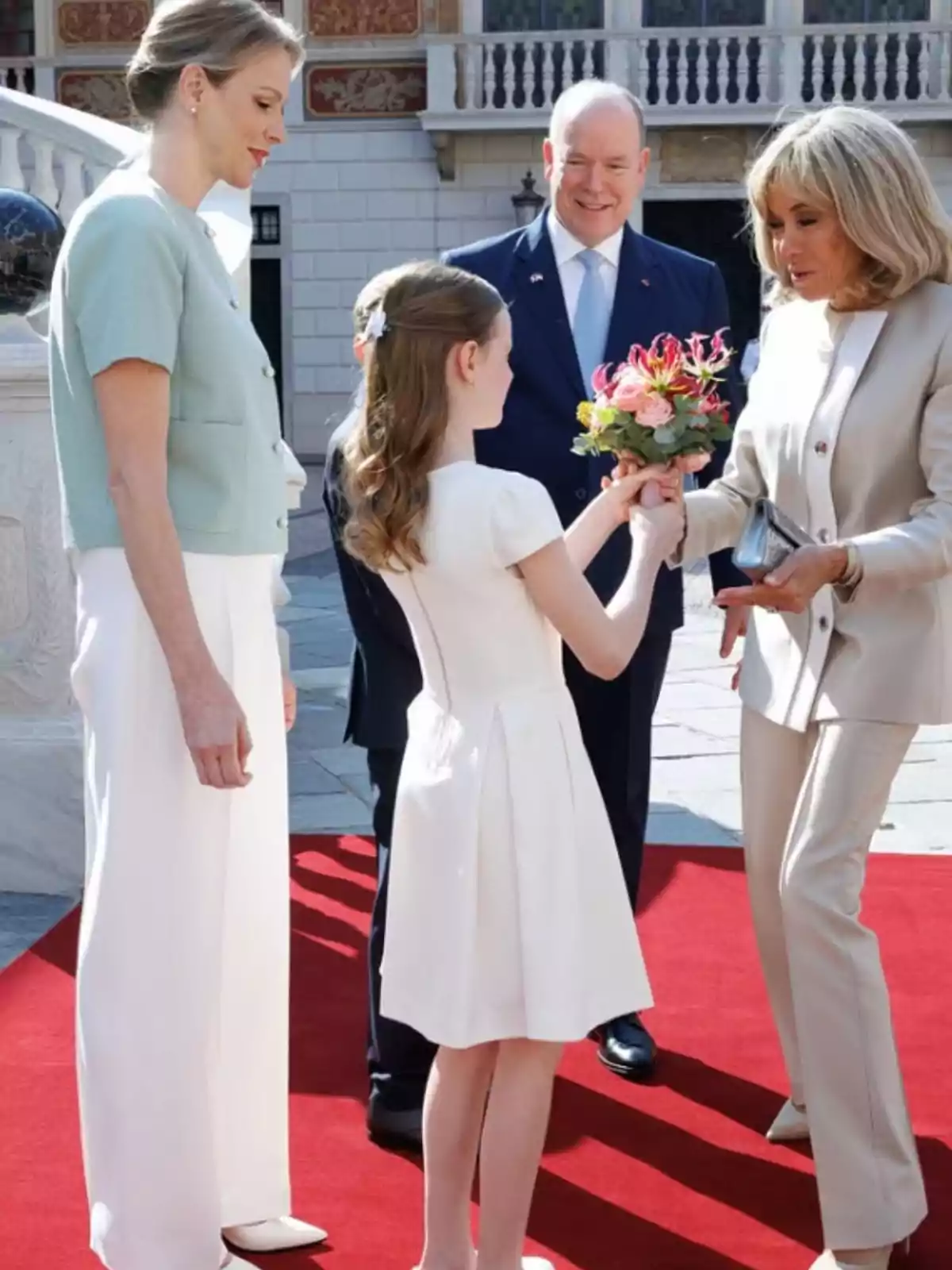 A girl hands a bouquet of flowers to an elegantly dressed blonde woman while two other people watch and smile on a red carpet in a formal setting. A girl hands a bouquet of flowers to an elegantly dressed blonde woman while two other people watch and smile on a red carpet in a formal setting.
