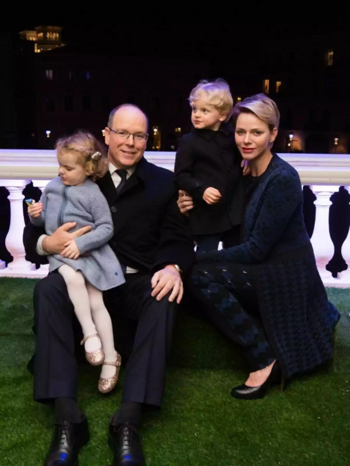 Albert of Monaco, Charlene and their children sitting on the grass at night, all dressed elegantly. Albert of Monaco, Charlene and their children sitting on the grass at night, all dressed elegantly.