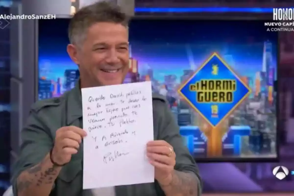 Alejandro Sanz somrient sosté un full de paper amb un missatge escrit a mà, mentre és en un plató de televisió amb el logotip d'"El Hormiguero" visible al fons.