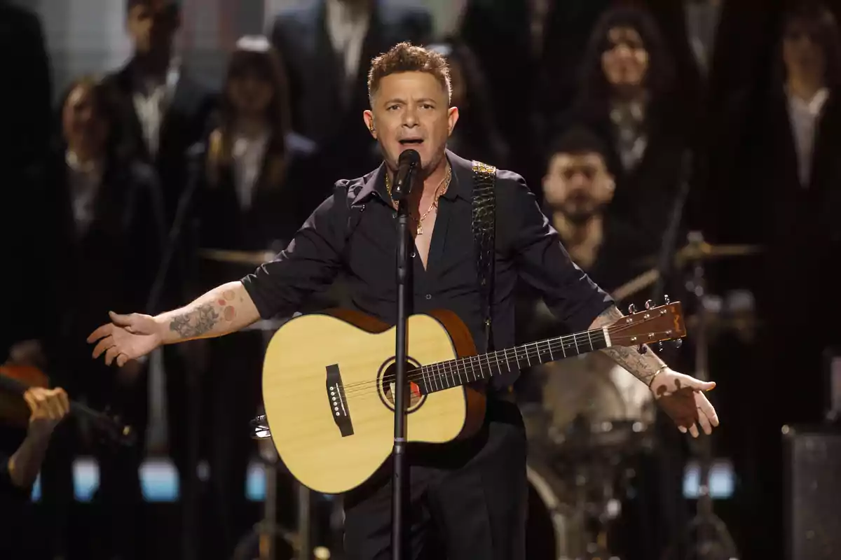 Alejandro Sanz singing and playing the guitar on a stage with musicians and backup singers in the background Alejandro Sanz singing and playing the guitar on a stage with musicians and backup singers in the background