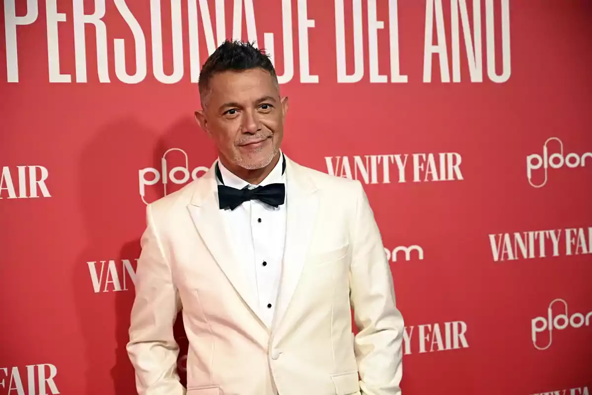Alejandro Sanz in a white tuxedo and black bow tie posing on a red carpet with a Vanity Fair backdrop Alejandro Sanz in a white tuxedo and black bow tie posing on a red carpet with a Vanity Fair backdrop