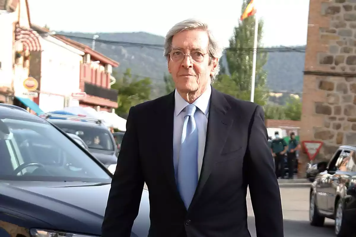 Un home gran amb vestit fosc i corbata blava caminant per un carrer amb edificis i cotxes al fons. Un home gran amb vestit fosc i corbata blava caminant per un carrer amb edificis i cotxes al fons.