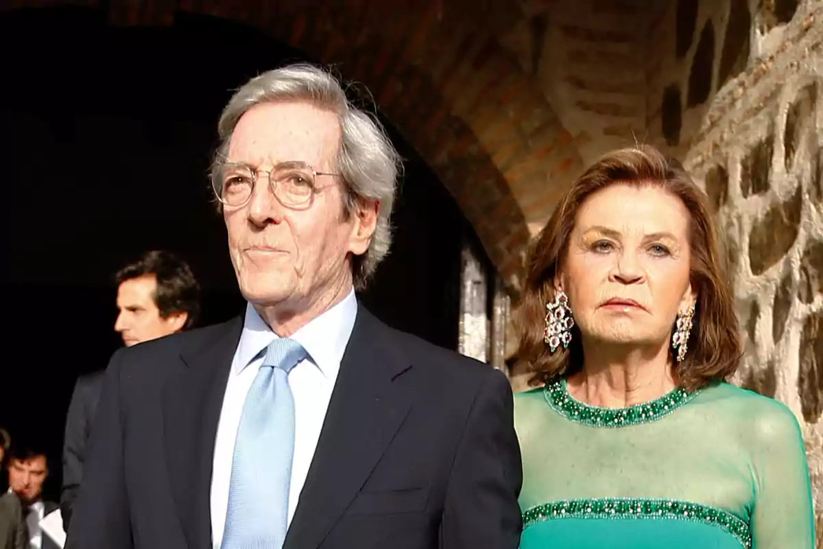 Una parella gran elegantment vestida, l'home porta vestit i corbata blau clar i la dona porta un vestit verd amb joies cridaneres, tots dos estan drets davant d'un fons de pedra. Una parella gran elegantment vestida, l'home porta vestit i corbata blau clar i la dona porta un vestit verd amb joies cridaneres, tots dos estan drets davant d'un fons de pedra.