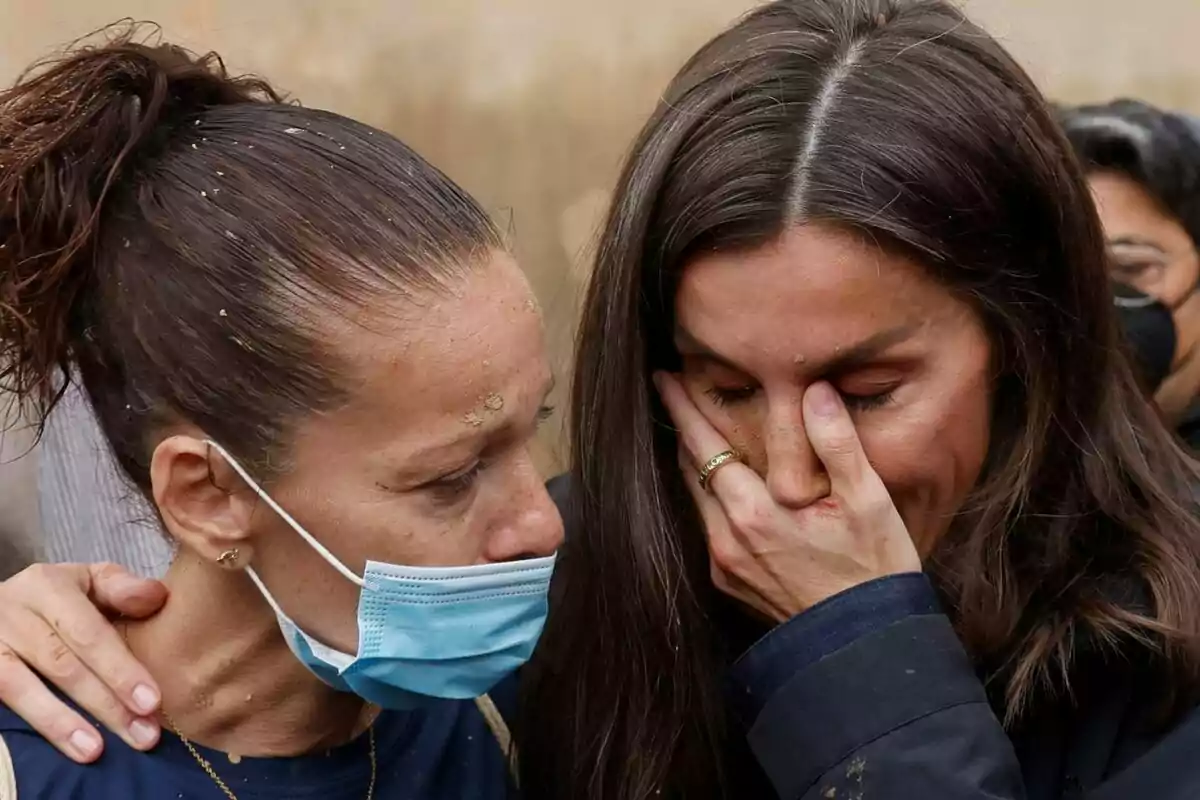 Letizia and a woman comfort each other, one of them wearing a mask and the other covering her face with her hand. Letizia and a woman comfort each other, one of them wearing a mask and the other covering her face with her hand.