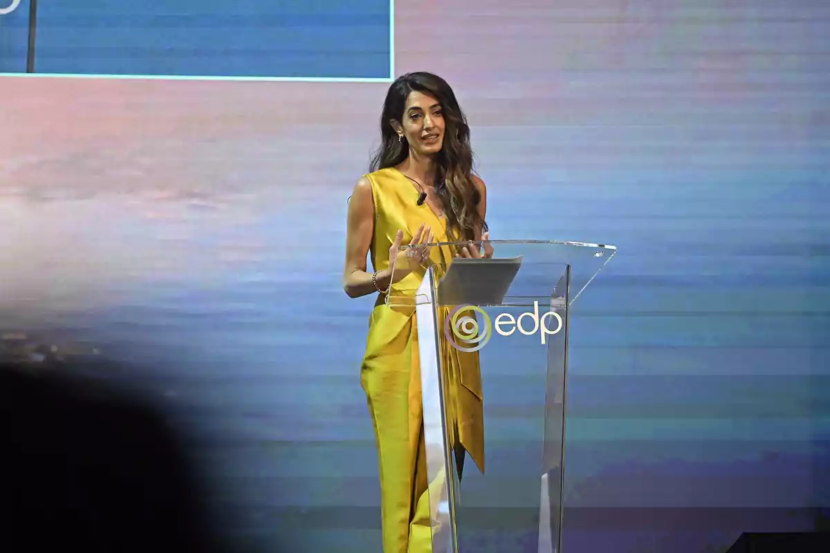 Amal Clooney in a yellow dress speaking at a transparent podium with the EDP logo at an event. Amal Clooney in a yellow dress speaking at a transparent podium with the EDP logo at an event.