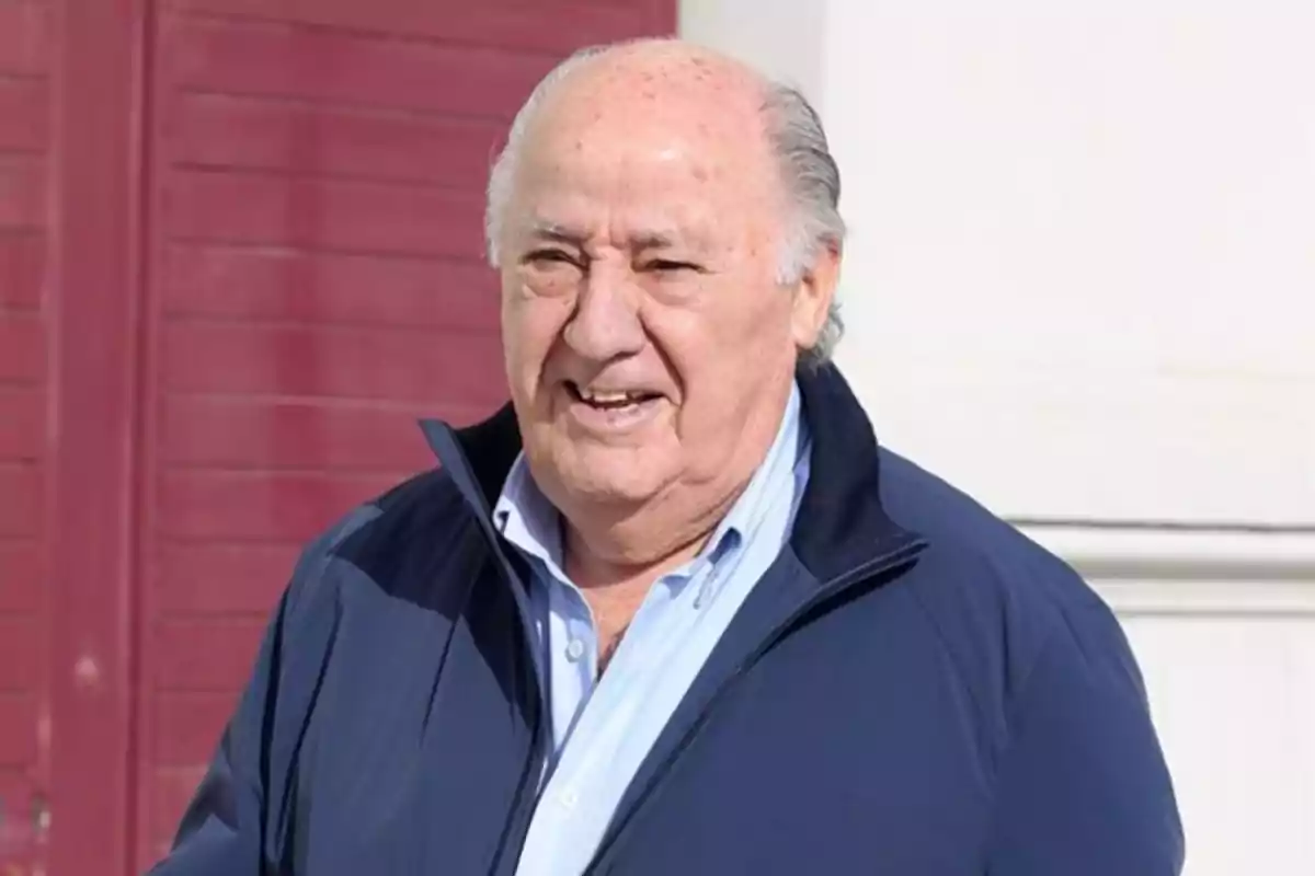 Amancio Ortega amb jaqueta blava i camisa clara somriu mentre està dret davant una paret de color vermell fosc.