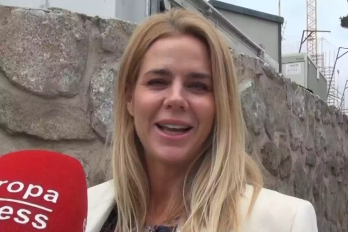 Amelia Bono somriu davant d'una paret de pedra mentre sosté un micròfon vermell. Amelia Bono somriu davant d'una paret de pedra mentre sosté un micròfon vermell.