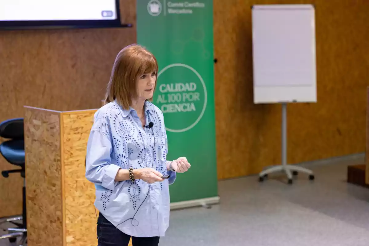 Una dona donant una presentació en un entorn professional amb un cartell verd que diu "Qualitat al 100 per Ciència" i un rodafoli al fons.