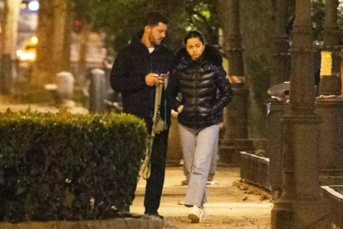 Una parella camina per un carrer arbrat a la nit, tots dos vesteixen roba d'abric i l'home sosté una corretja. Una parella camina per un carrer arbrat a la nit, tots dos vesteixen roba d'abric i l'home sosté una corretja.