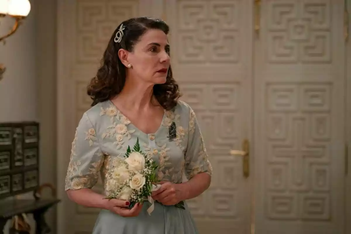 L'actriu Ana Fernández en una escena de 'Sueños de Libertad' amb un vestit blau clar brodat sostenint un petit ram de flors blanques en un interior elegant. L'actriu Ana Fernández en una escena de 'Sueños de Libertad' amb un vestit blau clar brodat sostenint un petit ram de flors blanques en un interior elegant.