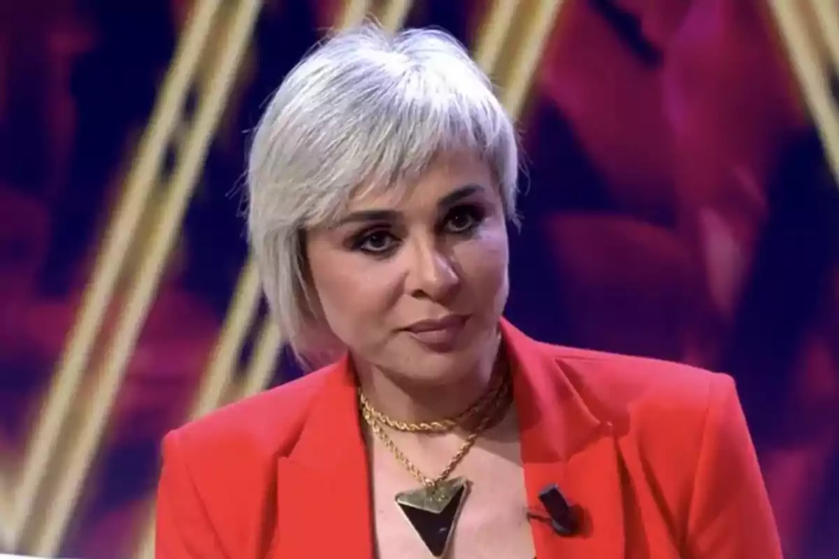 Ana María Aldón amb cabell curt i ros vestida amb un vestit vermell i un collaret daurat en un ambient il·luminat. Ana María Aldón amb cabell curt i ros vestida amb un vestit vermell i un collaret daurat en un ambient il·luminat.
