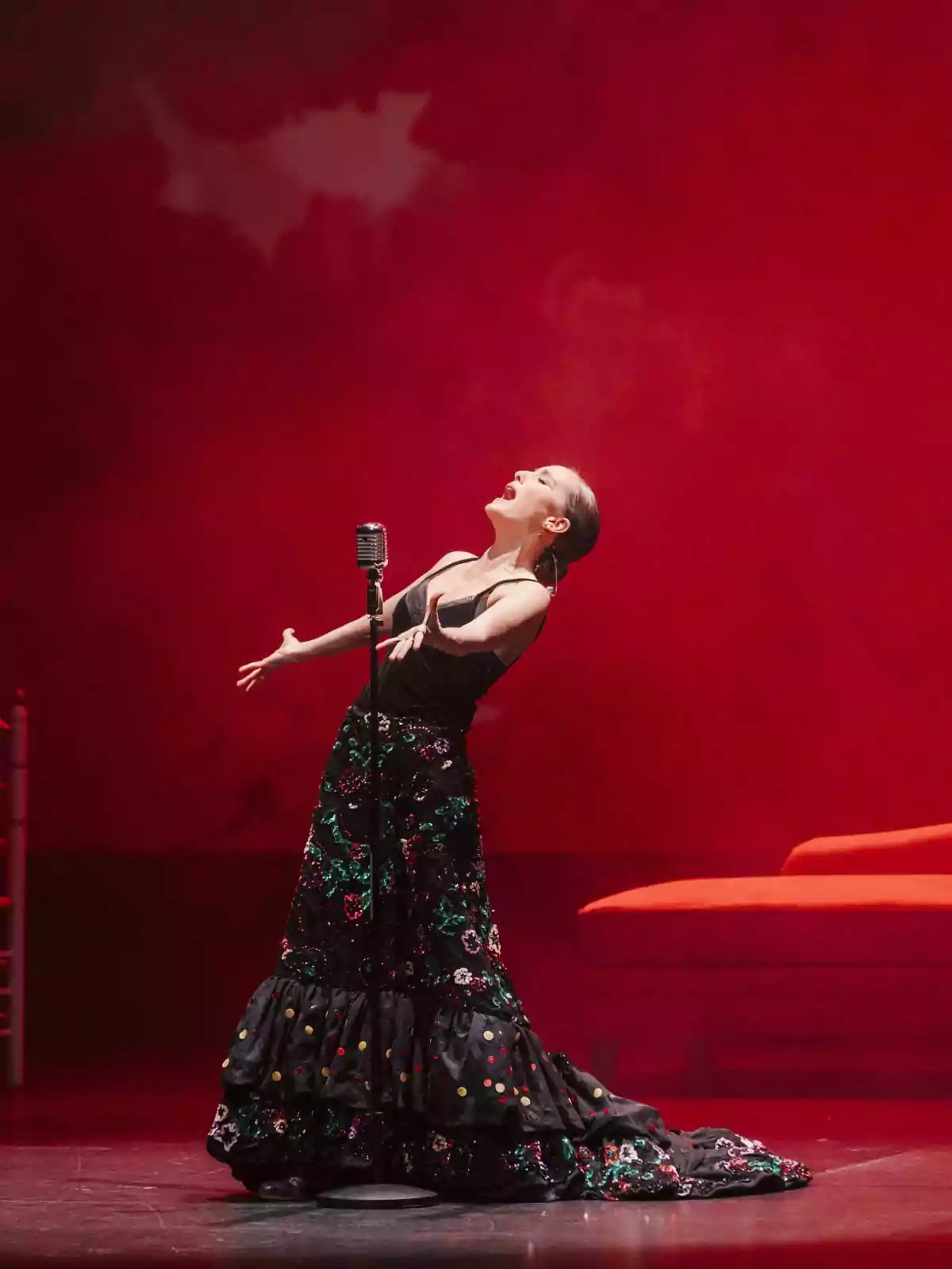 Anabel Dueñas canta apassionadament en un escenari amb fons vermell, usant un vestit llarg i colorit. Anabel Dueñas canta apassionadament en un escenari amb fons vermell, usant un vestit llarg i colorit.