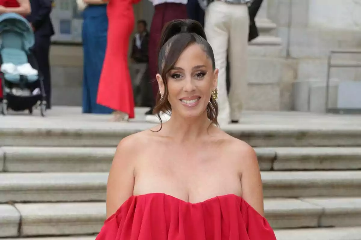 Anabel Pantoja con vestido rojo posando frente a unas escaleras con personas en el fondo.